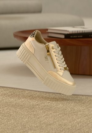 Beige plateau sneaker met gouden rits en accenten, witte veters, luipaardprint hiel, zwevend boven een beige tapijt met houten tafel en boeken op de achtergrond.