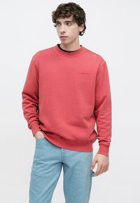 Κόκκινο φούτερ με ribbed λαιμόκοψη και μανίκια, με μικρό ανάγλυφο λογότυπο στο στήθος. Συνδυασμένο με ανοιχτό μπλε τζιν παντελόνι.