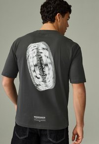 Tricou din bumbac gri, cu mâneci scurte, având un design grafic circular alb mare pe spate și o etichetă în partea de jos.