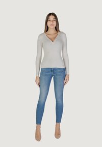 Maglia grigia chiara a maniche lunghe con scollatura a V e dettagli con bottoni, abbinata a jeans aderenti blu e tacchi nudi, creando un look casual.