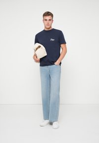 Camiseta de algodón azul marino con logo, combinada con jeans de mezclilla de pierna recta de color azul claro y zapatillas deportivas blancas; sosteniendo un bolso crema.