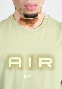 Ανοιχτό πράσινο βαμβακερό t-shirt με στρογγυλή λαιμόκοψη, μεγάλο σχέδιο κειμένου "AIR" και λογότυπο Nike σε μια διακριτική, αντίθετη απόχρωση.