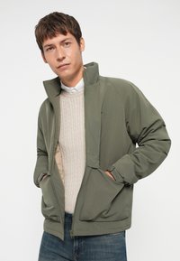 Chaqueta verde oliva con cremallera y alto cuello, puños elásticos y dos bolsillos frontales, superpuesta a un suéter de punto de cable en beige claro.