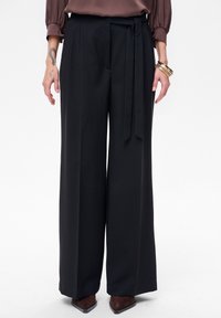 Czarne spodnie o szerokich nogawkach wykonane z gładkiego materiału. Z wiązaniem w pasie, plisami i płynną sylwetką. Noszone z brązowymi, szpiczastymi butami.