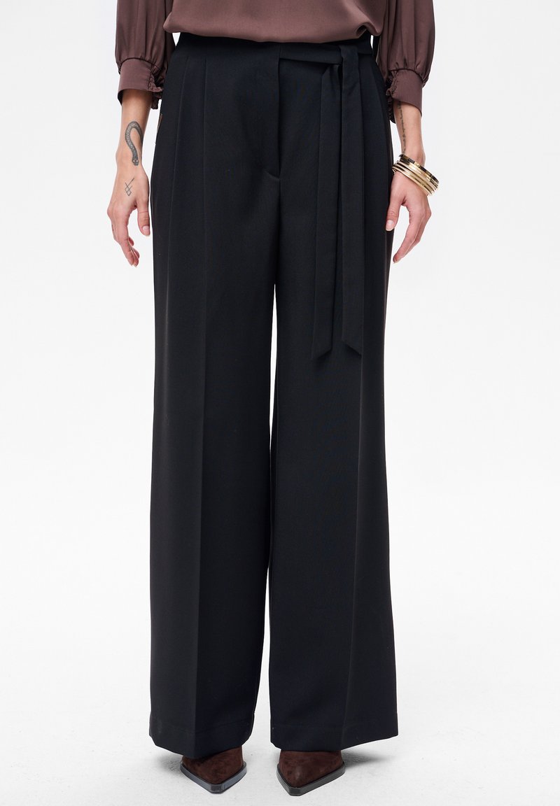 Czarne spodnie o szerokich nogawkach wykonane z gładkiego materiału. Z wiązaniem w pasie, plisami i płynną sylwetką. Noszone z brązowymi, szpiczastymi butami.