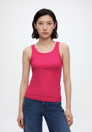 Γυναίκα με κοντά σκούρα μαλλιά που φοράει μαργαριταρένια σκουλαρίκια, φωτεινό ροζ αμάνικο τοπ και μπλε τζιν, στέκεται μπροστά σε απλό λευκό φόντο.