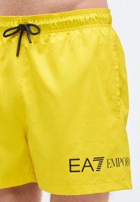 Dzelteni peldbikses no viegla materiāla; tām ir elastīga josta ar melnu auklu un melnā krāsā iespiests logotips.