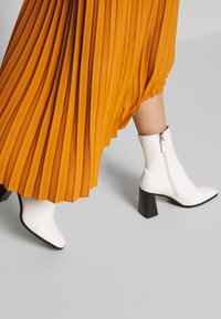 Bottines blanches à texture crocodile, à bout pointu et à talon carré, associées à une jupe orange fluide et plissée.