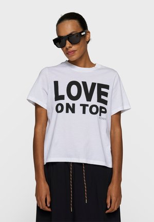 Donna con occhiali da sole oversize neri e una maglietta bianca con la scritta "LOVE ON TOP" in lettere nere grassetto.
