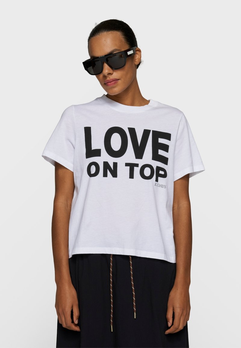 Femme portant des lunettes de soleil noires surdimensionnées et un T-shirt blanc avec l'inscription « LOVE ON TOP » en gros caractères noirs.