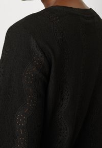 Maglione nero a trama con ritagli a scallop lungo le maniche. Il tessuto ha un aspetto a costine con una lieve lucentezza.