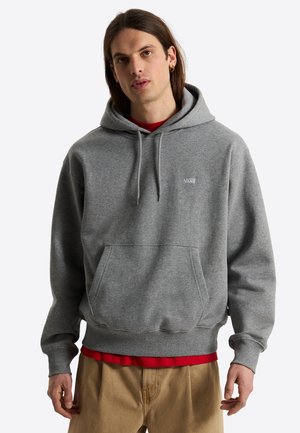Ung person med langt hår iført en grå Vans hættetrøje over en rød skjorte og beige bukser, stående mod en ensfarvet baggrund.