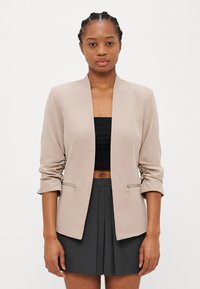 Bežový blazer s šálovým golierom, nariasenými rukávmi a striebornými zipsovými vrecami. Nosí sa s čiernym crop topom a šedou plisovanou sukňou.