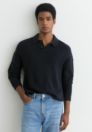 Jonge man die een zwart langarmig coltrui draagt en lichtblauwe spijkerbroek, staand tegen een witte muur met één hand in zijn zak.