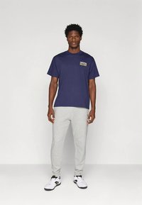 Άνδρας που στέκεται αντικριστά, φορώντας μπλε μπλούζα "Juventus", γκρι αθλητικά παντελόνια και λευκά αθλητικά παπούτσια σε μονόχρωμο φόντο.