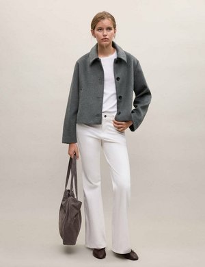 Femme portant une veste grise boutonnée, un pantalon blanc et des chaussures marron, tenant un sac fourre-tout gris sur fond neutre.