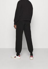 Sudadera y pantalones de chándal de algodón negro con puños elásticos, combinados con zapatillas blancas que tienen un logo rojo. Fondo liso.