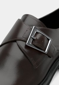 Chaussure habillée en cuir marron foncé avec une boucle rectangulaire argentée et une couture détaillée sur la lanière.