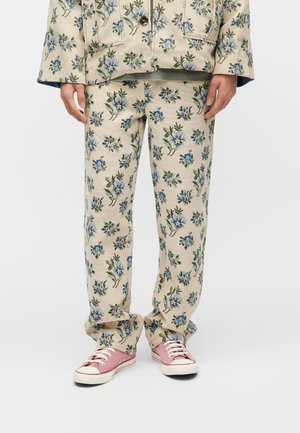 Henkilö, joka käyttää beigejä kukkakuosisia housuja ja vastaavaa takkia, yhdistettynä vaaleanpunaisiin kangastennareihin, seisomassa yksivärisen taustan edessä.