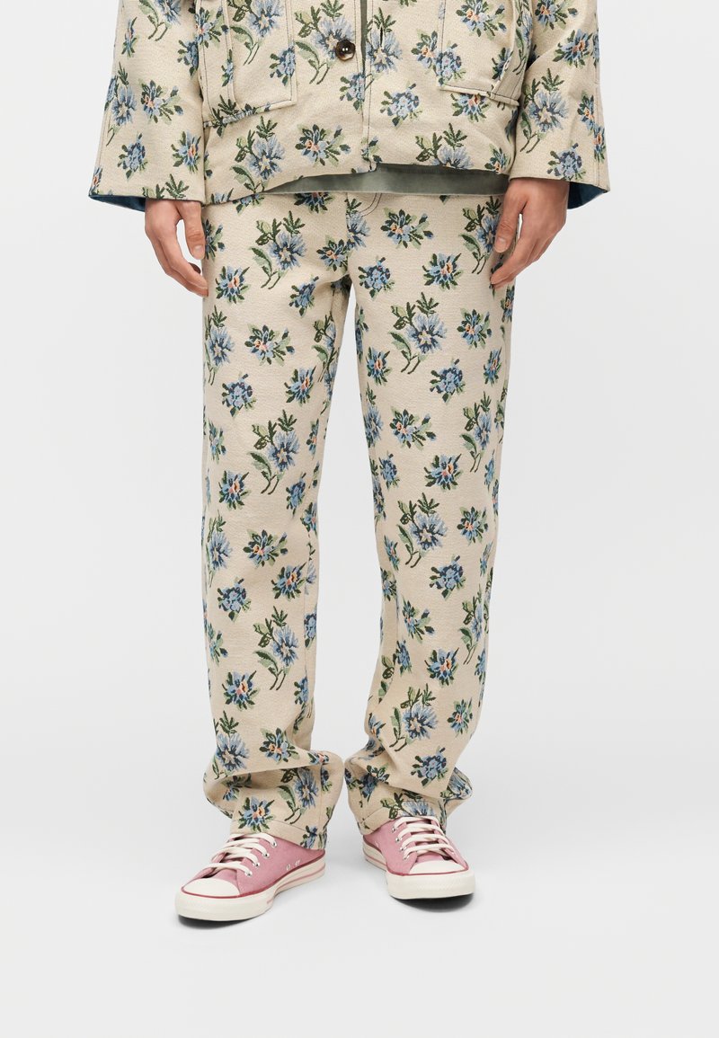 Άτομο που φοράει μπεζ φλοράλ παντελόνι και ταιριαστό μπουφάν, συνδυασμένα με ροζ πάνινα παπούτσια, στέκεται μπροστά σε απλή φόντο.