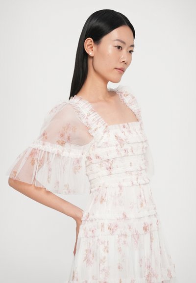 Biała sukienka w kwiaty z przezroczystymi bufkami i falbanami. Posiada kwadratowy dekolt oraz warstwowe falbany o miękkiej, lekkiej fakturze.