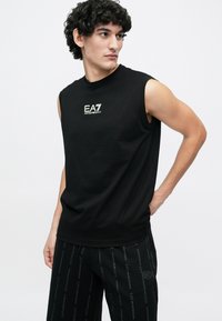 Νέος άνδρας με σγουρά μαλλιά, φορώντας μαύρη αμάνικη μπλούζα EA7 Emporio Armani και μαύρο παντελόνι με σχέδια, στέκεται με το ένα χέρι στη μέση.