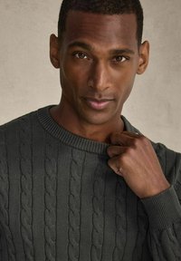 Gebreide trui in donkergroen met een kabelpatroon, voorzien van een ribgebreide ronde hals en manchetten. De hand van het model rust op de kraag.