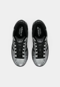 Melnas un sudrabotas Adidas kedas ar teksturētu apvalka purngalu, trim melnām svītrām sānos un melnām šņorēm. Bez ievērojamām rakstām vai akcentiem.