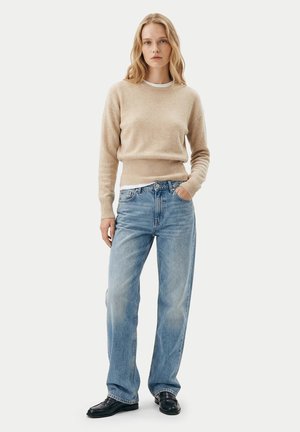 Frau steht und trägt einen beigen Langarmpullover, hellblaue hoch taillierte Jeans und schwarze Loafers vor einem einfarbigen hellen Hintergrund.