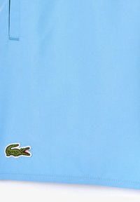 Jasnoniebieskie materiałowe szorty z zielonym haftowanym logo krokodyla na dolnym brzegu. Gładka tekstura z delikatnymi detalami szwów.