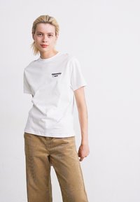 Junge Person mit kurzen blonden Haaren, die ein weißes T-Shirt mit Grafik und leicht braune Cordhosen trägt, vor einem einfarbigen weißen Hintergrund.