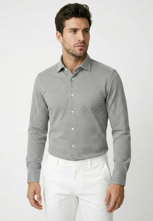 Man in een nauwsluitend zwart-wit geruit shirt met lange mouwen en witte broek, staand tegen een effen lichte achtergrond.