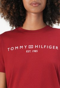 Raudona medvilninė marškinė su apvalia iškirpte ir trumpomis rankovėmis, su balta tekstine logo "TOMMY HILFIGER EST. 1985" ant krūtinės.