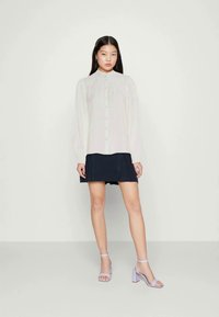 Blusa blanca con botones en la parte delantera y detalles plisados, combinada con una falda azul marino. El modelo lleva sandalias de tacón alto lavanda sobre un fondo neutro.