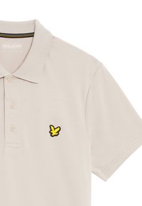 Lichtbeige polo shirt met een klassieke kraag en een drieknoopsluiting. Heeft een geel en zwart geborduurd logo op de linkerborst.