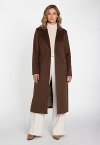 Brun lang frakke med hakket revers, knaplukning foran og to lommer. Foret med leopardmønster. Båret over en cremefarvet rullekrave og bukser.