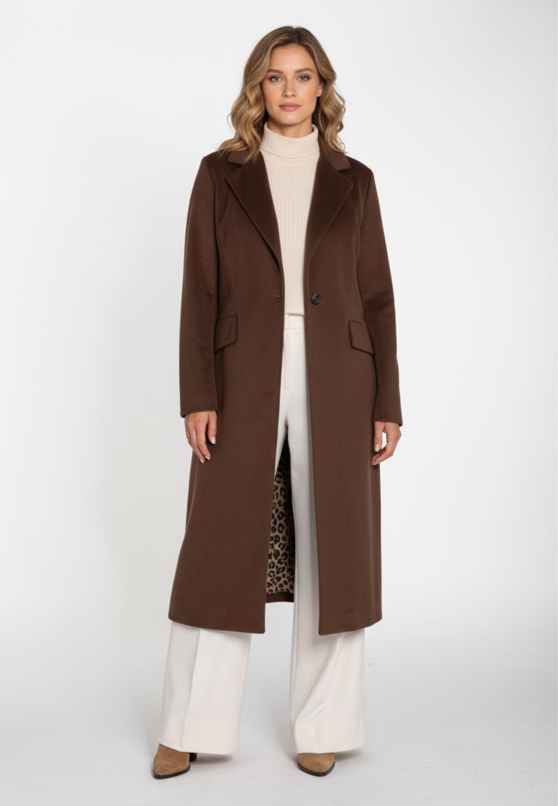 Brun lang frakke med hakket revers, knaplukning foran og to lommer. Foret med leopardmønster. Båret over en cremefarvet rullekrave og bukser.