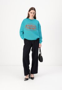 Sweatshirt turquesa com gráfico "AMERICAN VINTAGE"; combinado com calças largas de risca de giz pretas e sapatos de salto alto pretos. Mala preta.