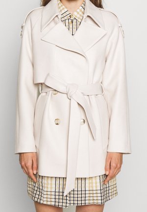 Femme portant un manteau crème ceinturé à double boutonnage sur une robe à carreaux beige et blanc, debout devant un fond uni.