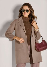 Frau in beigem Mantel, braunem Pullover, Sonnenbrille und mit einer burgunderfarbenen Handtasche mit goldenen Nieten, die gegen eine helle Wand steht.