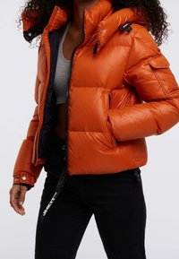 Pomarańczowa ocieplana kurtka na zamek błyskawiczny, z wysokim kołnierzem i bocznymi kieszeniami. Z czarnymi akcentami oraz zatrzaskami na mankietach.