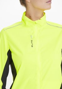 Hochsichtbare gelbe Sportjacke mit schwarzen Seitenpaneelen; verfügt über einen Stehkragen und reflektierende Akzente für bessere Sichtbarkeit.