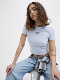 Lyseblå kortermet crop top med teksten "DUMP HIM" i svart. Bæres sammen med lyse jeans og en grå rutete skjorte knyttet rundt midjen.