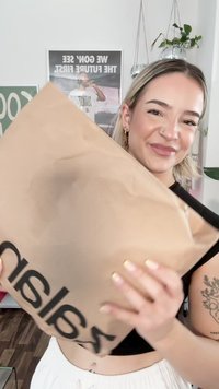 Mlada žena s kratkom plavom kosom se smiješi, držeći veliki smeđi paket iz Zalanda u modernoj prostoriji s posterima i biljkama u pozadini.