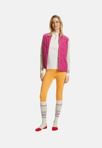 Model trägt eine rosa gesteppte Weste über einem weißen Oberteil, orangefarbene Leggings und knielange Socken mit mehrfarbigen Streifen und roten Akzenten.
