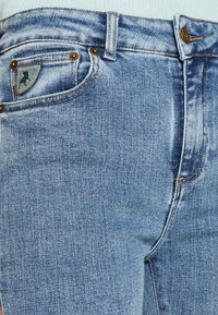 Svijetloplave traperice s izblijedjelim završetkom, koje imaju prednju torbicu s plavim i sivim zakrpanom, bakrene zakovice i kopčanje na dugme.
