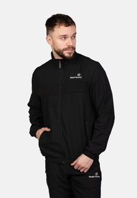 Schwarze Zip-Jacke mit hohem Kragen, die ein horizontales Panel über der Brust und ein besticktes Logo aufweist. Weiches, glattes Material.