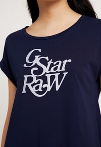 Tmavě modré bavlněné tričko s velkým bílým stylizovaným logem "G-Star RAW" uprostřed. Krátké rukávy a volný střih.