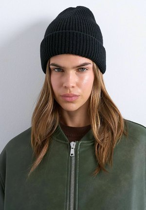 Woman with long hair wearing a black knit beanie, green zip-up jacket, and brown top against a plain light background.