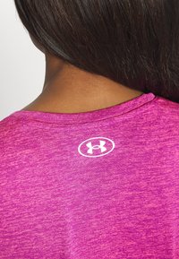 Mujer con cabello oscuro que lleva una camiseta deportiva de color rosa jaspeado, mostrando un logo blanco de Under Armour centrado en la parte superior de la espalda.